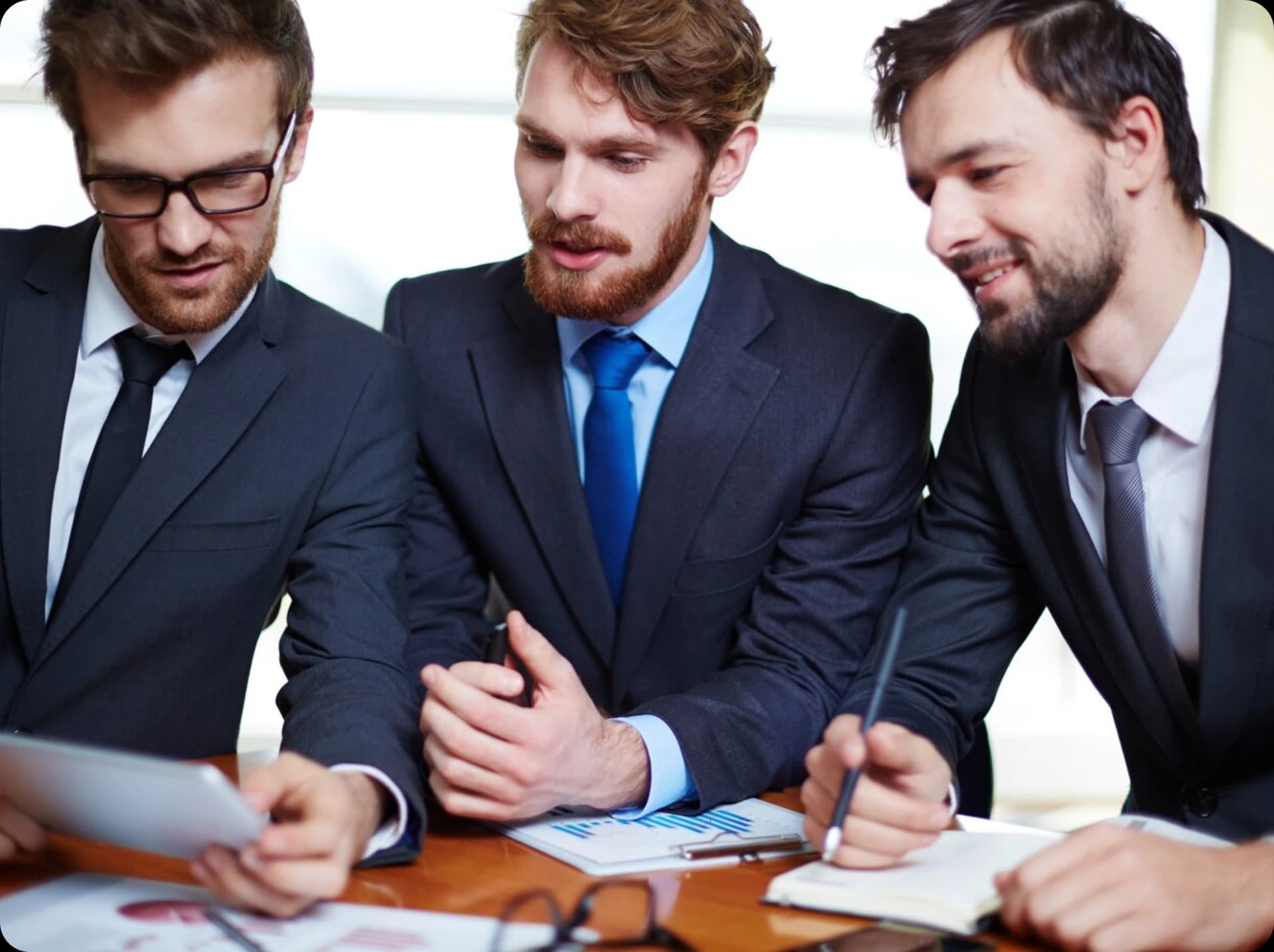 Three businessmen in suits engaged in a discussion, looking at a tablet and documents on the table, in a professional office environment.